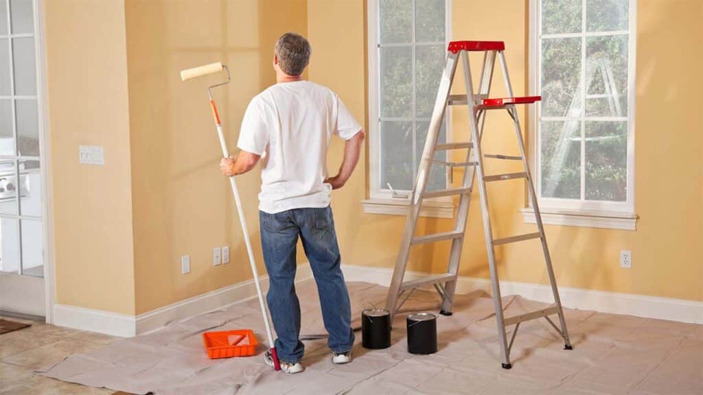A man paints a room with yellow walls, ladder, and supplies ready