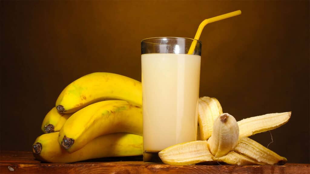 A glass of banana juice with a yellow straw is placed beside a bunch of ripe bananas and a peeled banana on a wooden surface