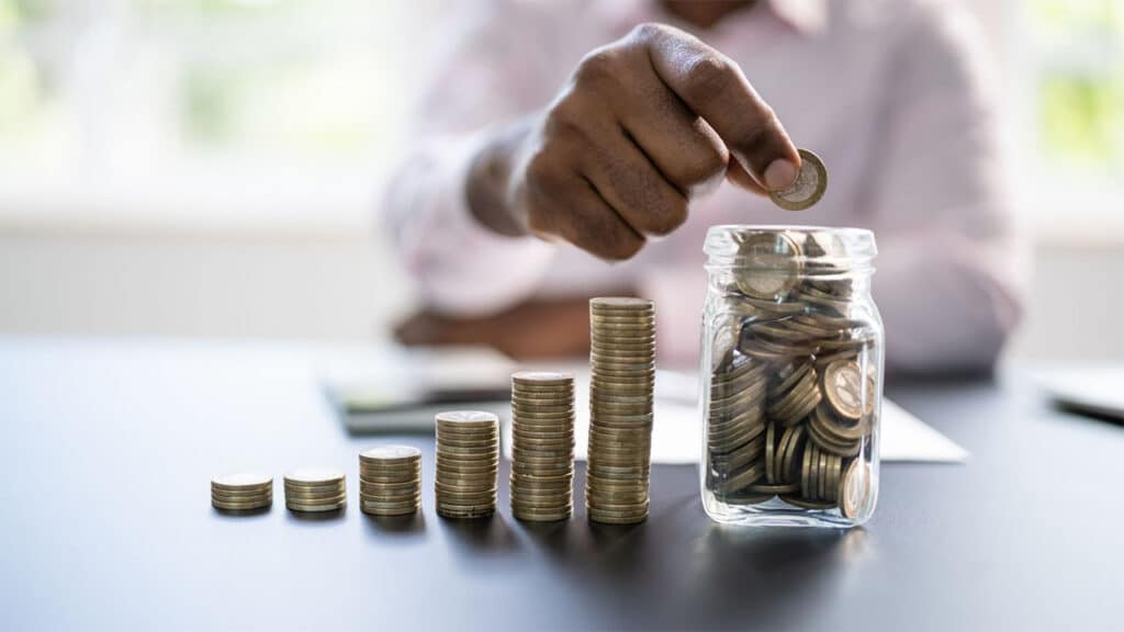 Person stacking coins and placing them into a glass jar, symbolizing savings and financial growth
