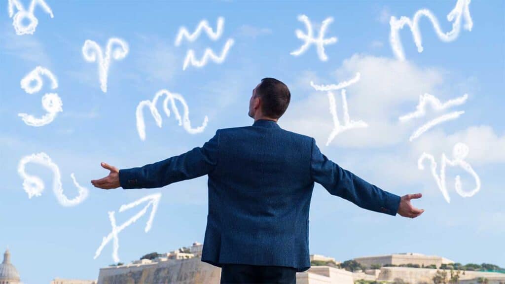 A man stands with arms outstretched, facing zodiac signs in the sky